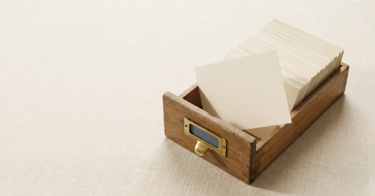 A wooden card-index drawer on a warm linen surface, packed with cream archival cards and a single card tilted forward, a physical metaphor for progressive disclosure